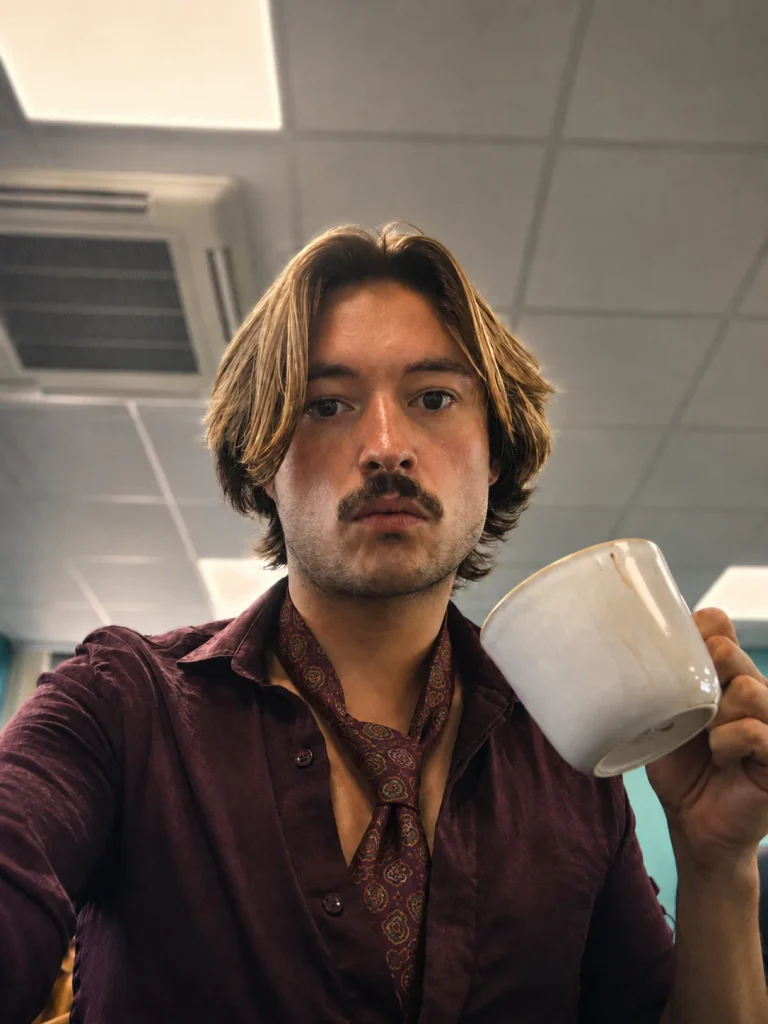 Seated office close-up showing a plum shirt with an open collar, patterned tie and a white mug.