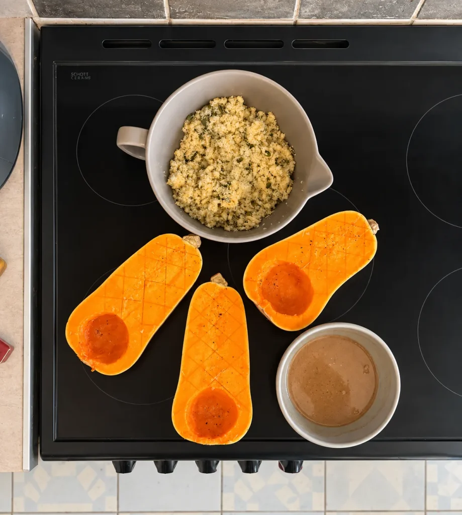 Three scored butternut squash halves showing their small natural seed cavities, with the topping and seasoning mixture ready beside them.