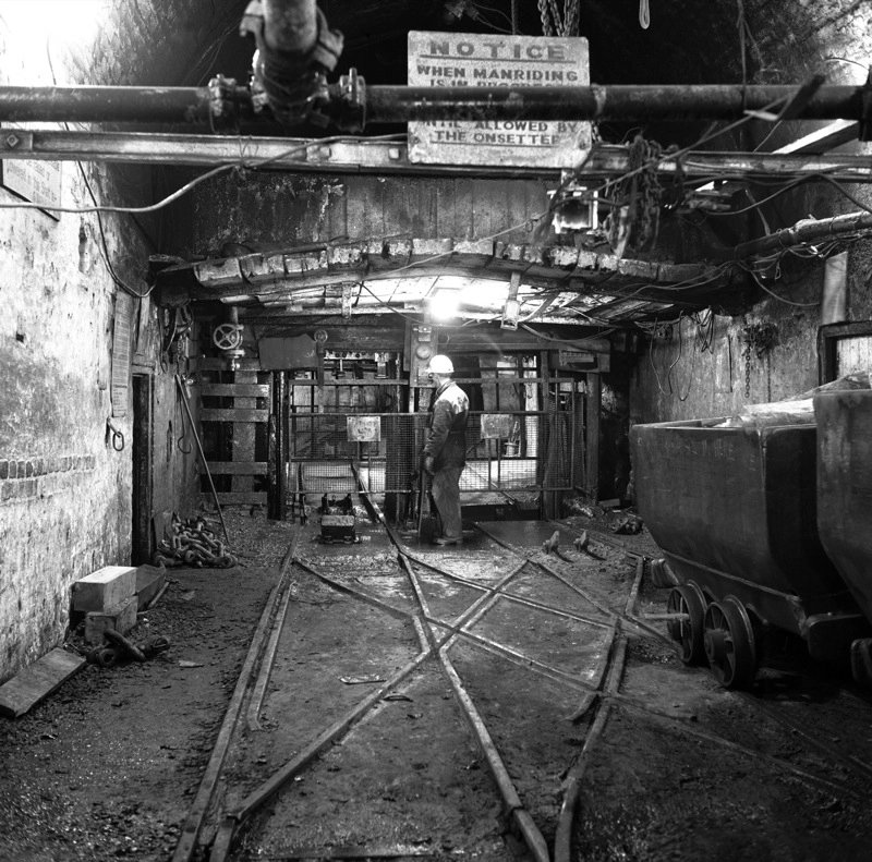 Black-and-white photograph of the pit bottom at Cwmtillery Colliery in Wales in 1977, showing a miner standing among underground rail tracks, pipes and coal tubs beneath a low mine roof.