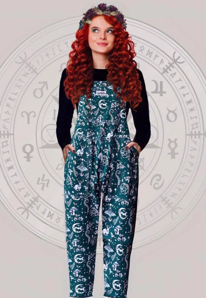 Person wearing Jambats Spirit of Magick dungarees printed with moons, mushrooms, moths and other witchy motifs over a black long-sleeved top.