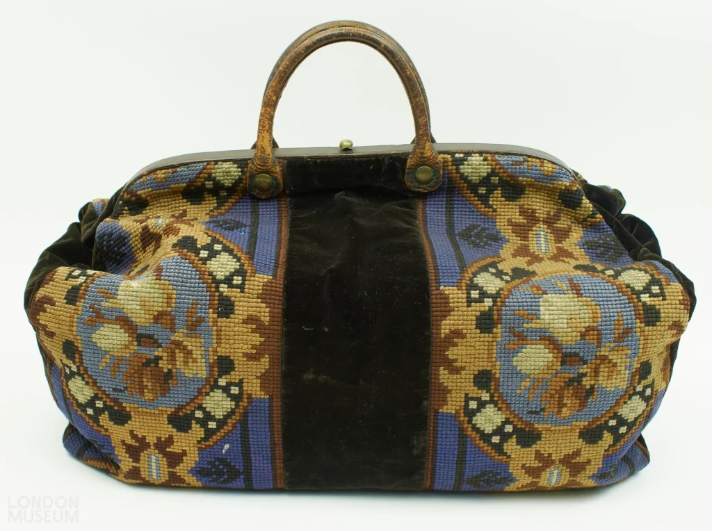 Victorian carpetbag with blue, brown and beige woolwork, dark velvet panels and leather handles, dated 1851–1860.